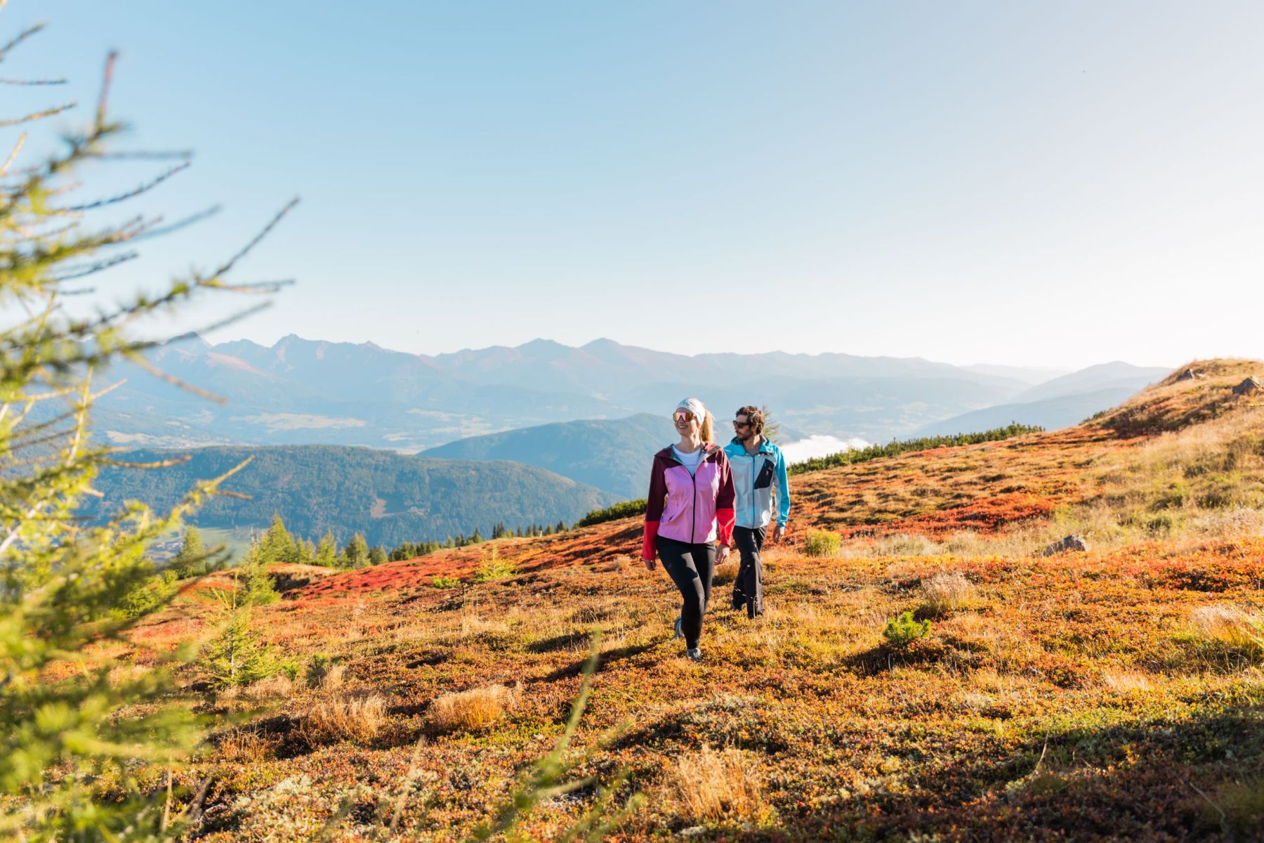 Zwei Personen wandern auf einem herbstlichen Bergpfad im Salzburger Lungau, mit Blick auf bewaldete Hügel und Berge im Hintergrund.
