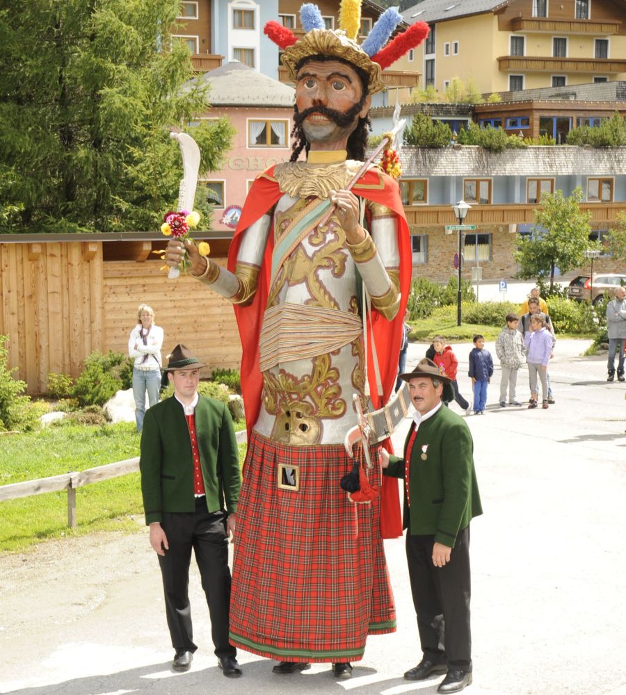 Große Figur in traditioneller Kleidung mit buntem Kopfschmuck, flankiert von zwei Männern in grünen Trachtenjacken.
