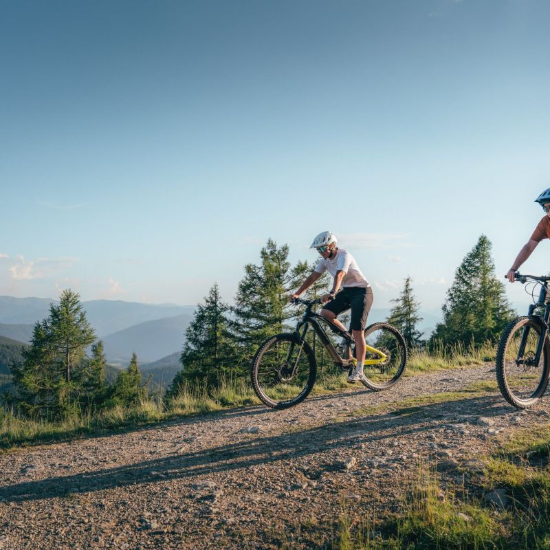 Zwei Personen fahren mit Mountainbikes auf einem Schotterweg durch eine bergige Waldlandschaft im Salzburger Lungau.