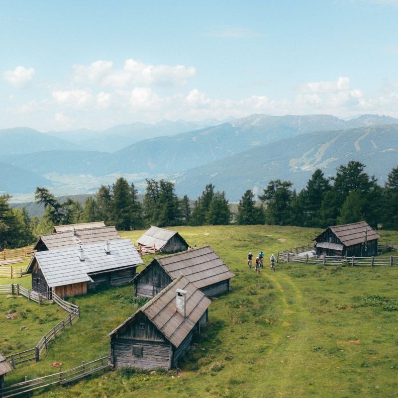 Mehrere Holzhütten in grüner Berglandschaft, umgeben von Bäumen, mit Blick auf entfernte Berge unter blauem Himmel.