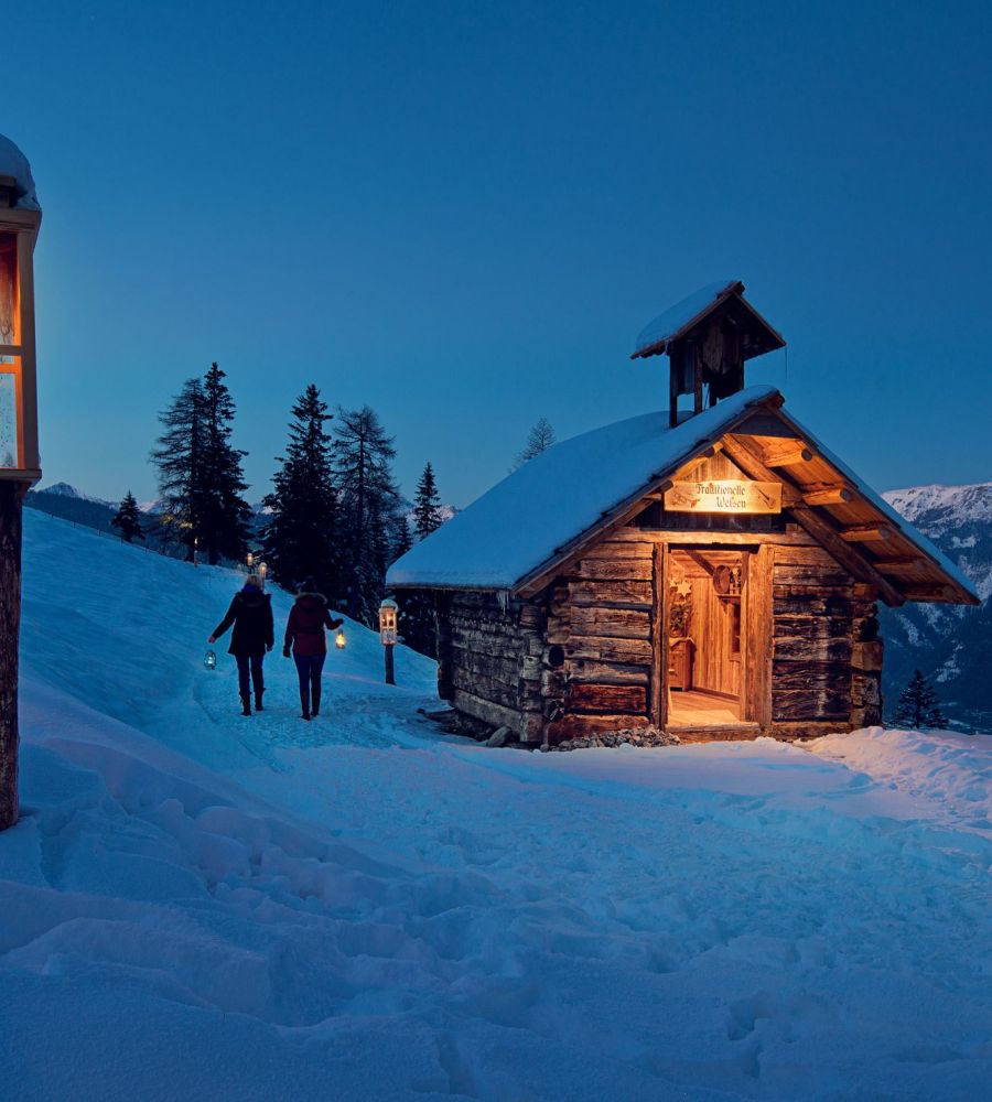 Ein verschneiter Hang im Salzburger Lungau bei Nacht. Eine beleuchtete Hütte und Laterne, Personen tragen Laternen.