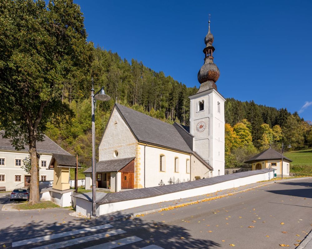 Pfarrkirche Thomatal mit Zwiebelturm vor bewaldetem Hügel, blauer Himmel, Straße und Baum im Vordergrund, Herbstfarben.