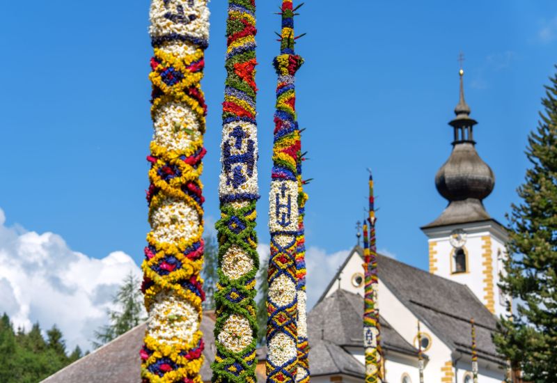 Bunte, verzierte Stangen vor einer Kirche mit Zwiebelturm, blauer Himmel im Hintergrund, Salzburger Lungau.