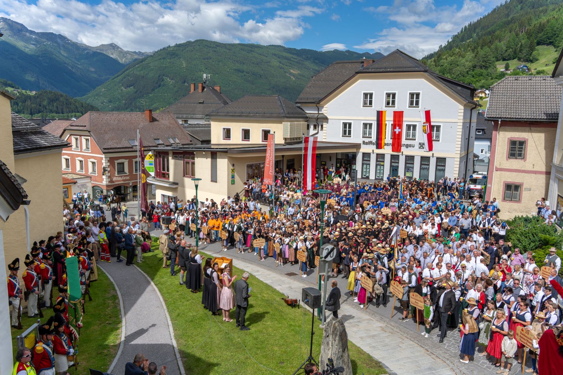 Eine große Menschenmenge in traditioneller Kleidung versammelt sich auf einem Platz in einem malerischen Dorf mit Bergkulisse.