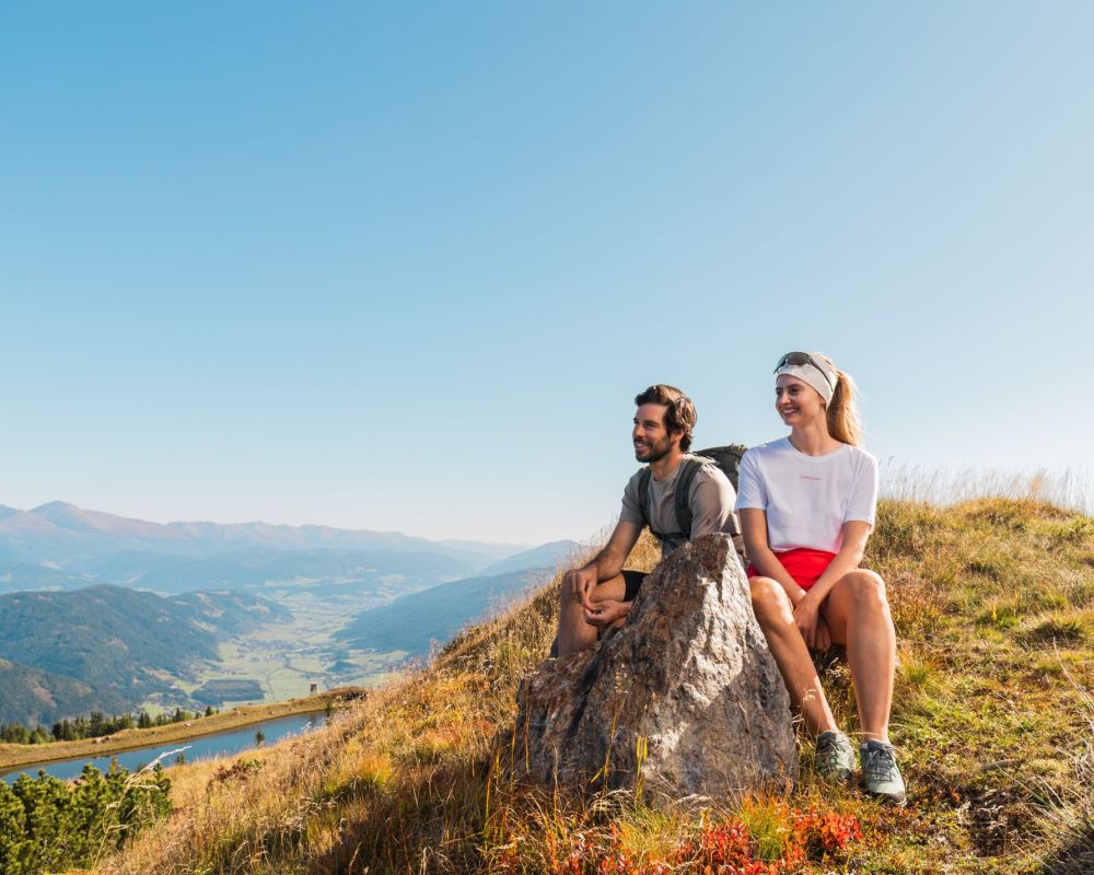 Zwei Personen sitzen auf einem Hügel im Salzburger Lungau, mit Blick auf Berge und Tal bei klarem Himmel.