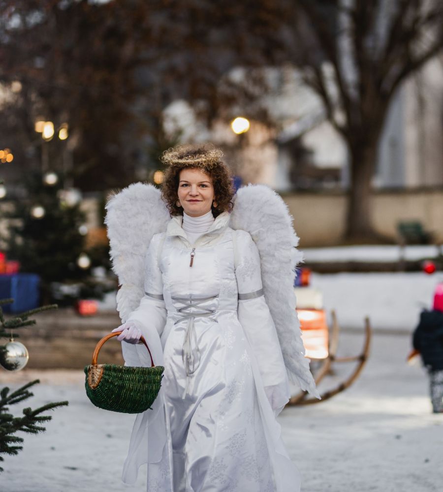 Ein Engel in weißem Kostüm mit Flügeln und Korb geht durch einen verschneiten Weihnachtsmarkt mit Tannenbäumen.