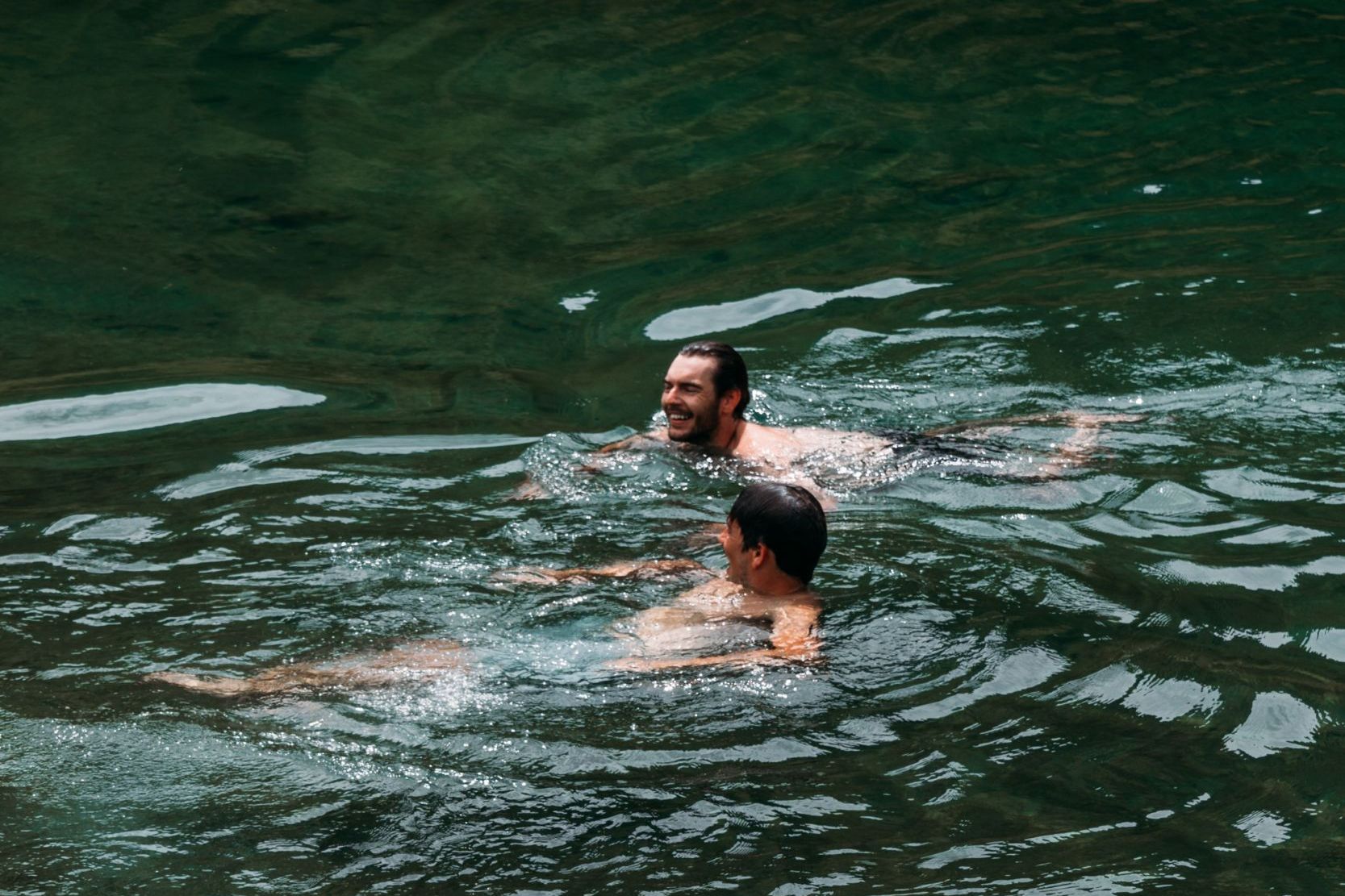 Zwei Personen schwimmen in einem klaren, grünen See. Die Wasseroberfläche reflektiert das Licht.