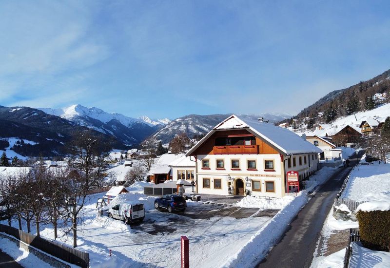 Schneebedeckte Landschaft im Salzburger Lungau, großes Gebäude mit "Metzgerstubn" Schild, umgeben von Bergen und klarer Himmel.