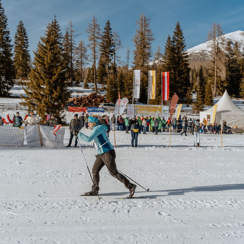 Langläufer auf verschneiter Strecke im Salzburger Lungau, Zuschauer und Zelte im Hintergrund, umgeben von Nadelbäumen.