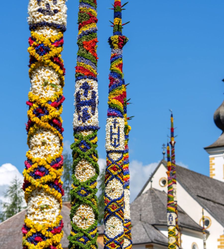 Bunte, verzierte Stangen vor einer Kirche mit Zwiebelturm, blauer Himmel im Hintergrund, Salzburger Lungau.