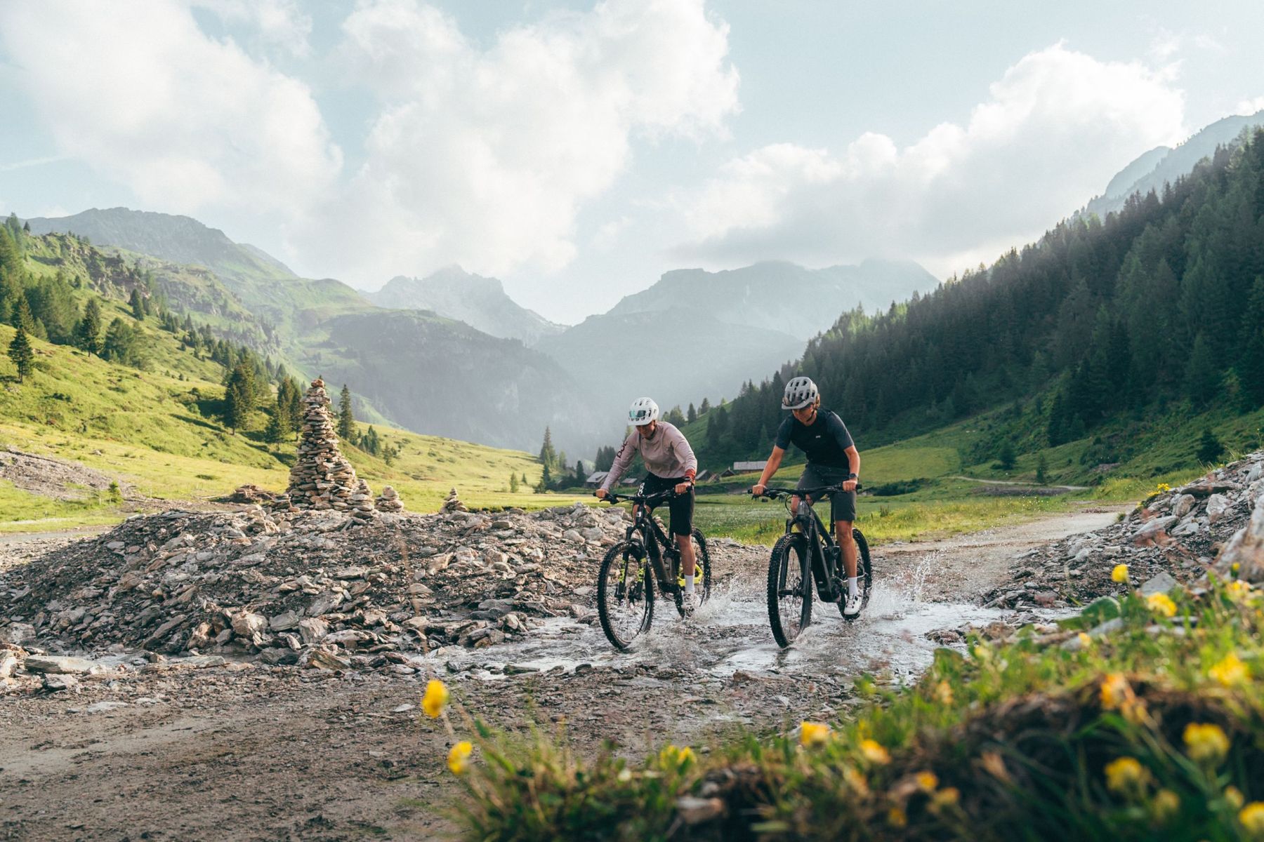 Zwei Radfahrer fahren durch eine Pfütze auf einem Bergweg im Salzburger Lungau, umgeben von grünen Hügeln und Bergen.