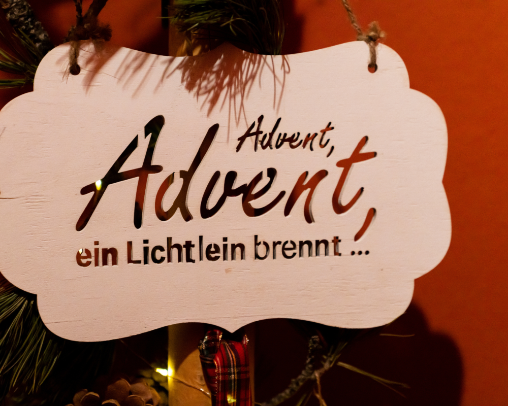 Holzschild mit dem Text "Advent, Advent, ein Lichtlein brennt..." vor einem roten Hintergrund, dekoriert mit Tannenzweigen.
