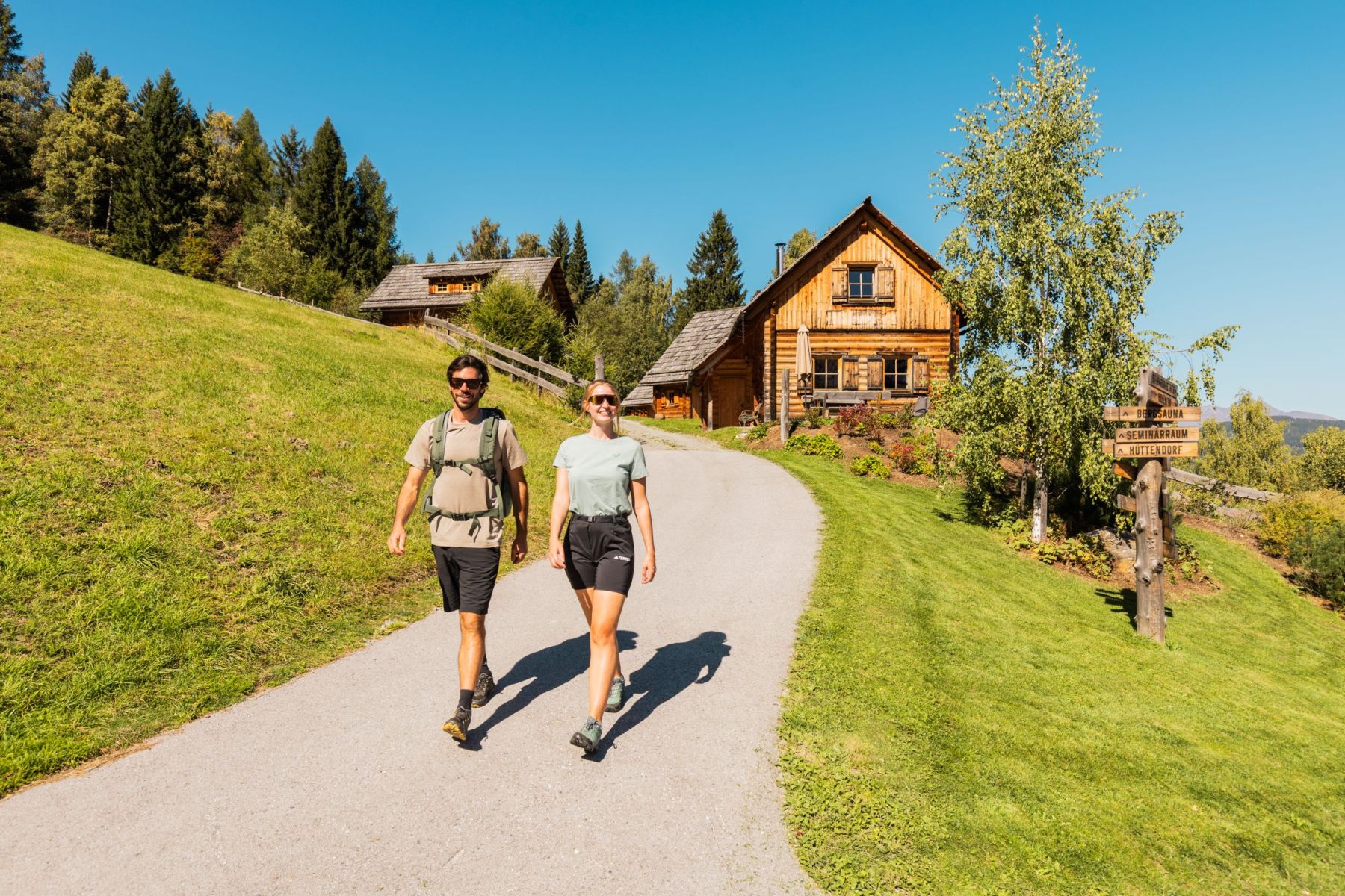 Zwei Personen wandern auf einem Weg neben einer Holzhütte im Salzburger Lungau, umgeben von Wiesen und Bäumen.