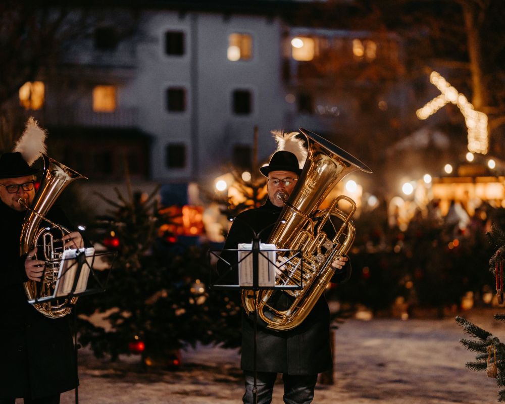 Zwei Musiker mit Blechblasinstrumenten spielen bei einer festlichen Veranstaltung im Freien, umgeben von Lichtern und Tannen.