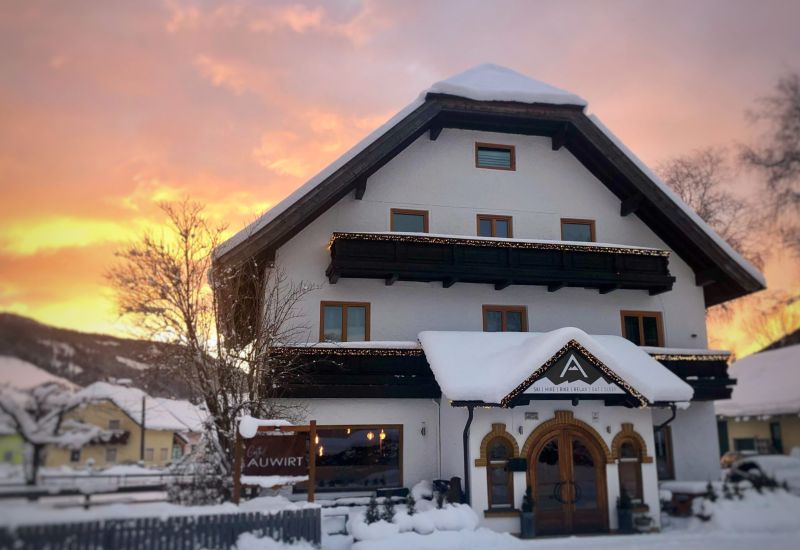 Zweistöckiges Haus im Schnee bei Sonnenuntergang, Schild "Auwirt" vorne, verschneite Berge im Hintergrund.