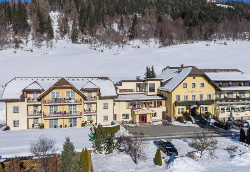 Gelbes Hotel im Salzburger Lungau, umgeben von verschneiten Feldern und Wald, unter klarem, blauem Himmel. Winterliche Szene.