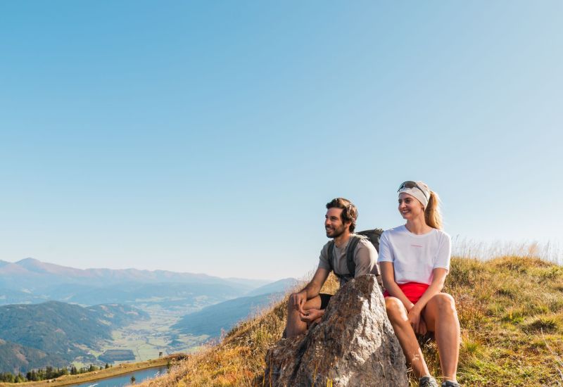 Zwei Personen sitzen auf einem Hügel im Salzburger Lungau, mit Blick auf Berge und Tal bei klarem Himmel.