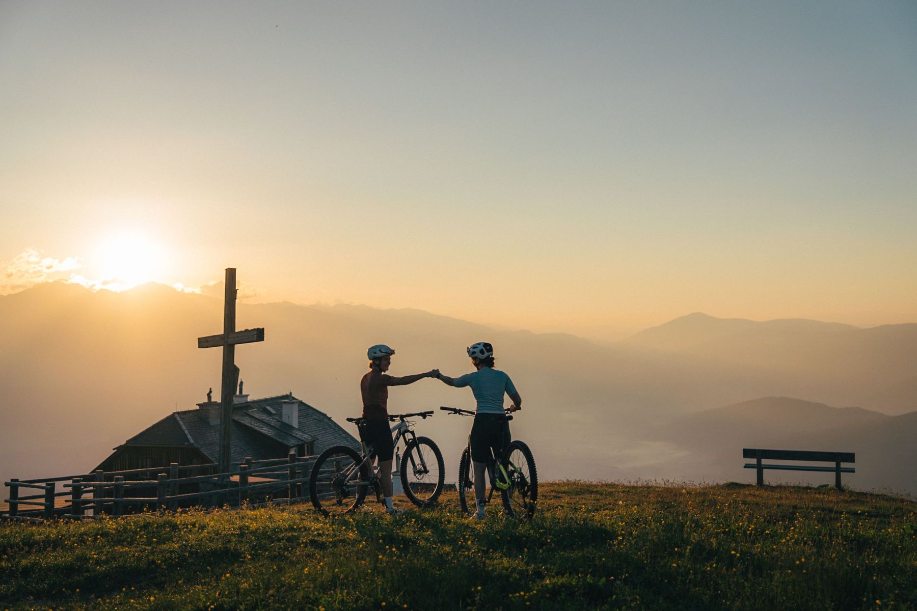 Zwei Radfahrer geben sich bei Sonnenuntergang auf einem Hügel im Salzburger Lungau die Hand, neben einem Holzkreuz.