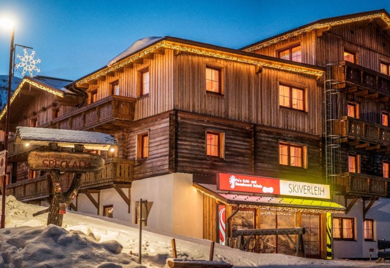 Almhütte bei Dämmerung im Salzburger Lungau, beleuchtet mit Lichtern, umgeben von Schnee. Schild mit der Aufschrift "Speckalm".