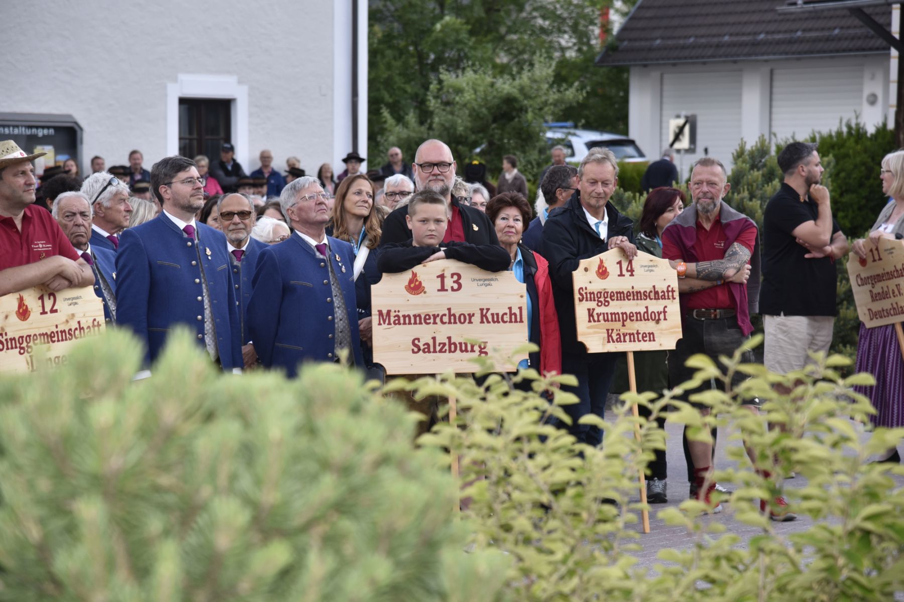 Eine Gruppe von Menschen steht draußen mit Schildern für Männerchor Kuchl Salzburg und Singgemeinschaft Krumpendorf Kärnten.