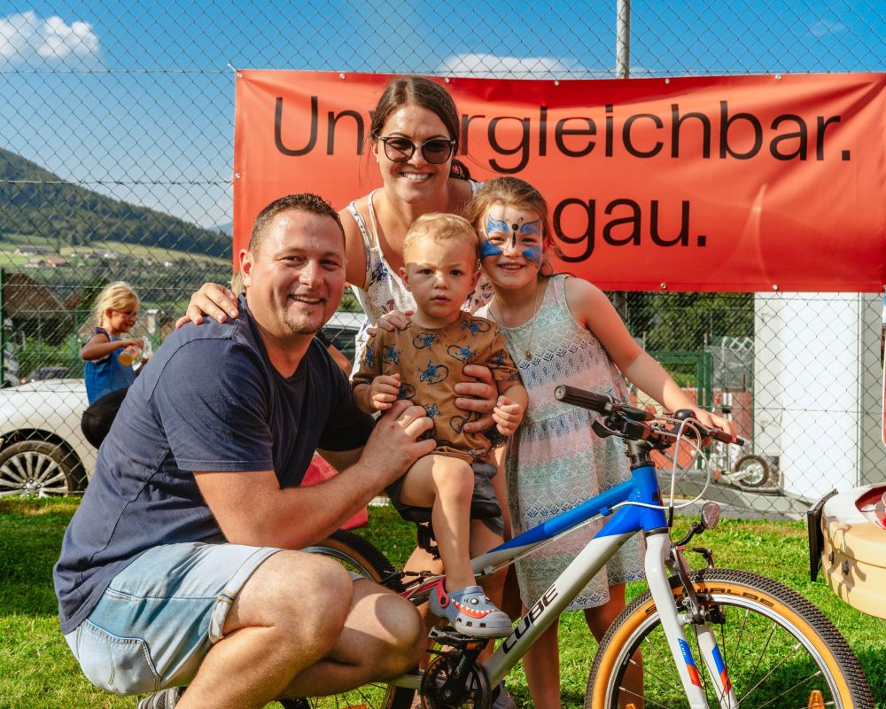 Familie vor einem Schild mit "Unvergleichbar. Lungau." im Hintergrund. Ein Kind mit Gesichtsbemalung steht neben einem Fahrrad.