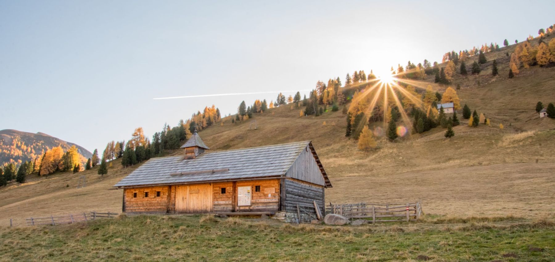 Holzhütte mit Schindeldach auf Bergwiese, Sonnenaufgang hinter Berghang. Klare, helle Atmosphäre im Salzburger Lungau.