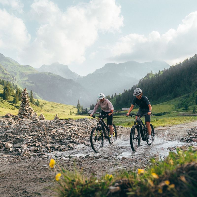 Zwei Radfahrer fahren durch eine Pfütze auf einem Bergweg im Salzburger Lungau, umgeben von grünen Hügeln und Bergen.