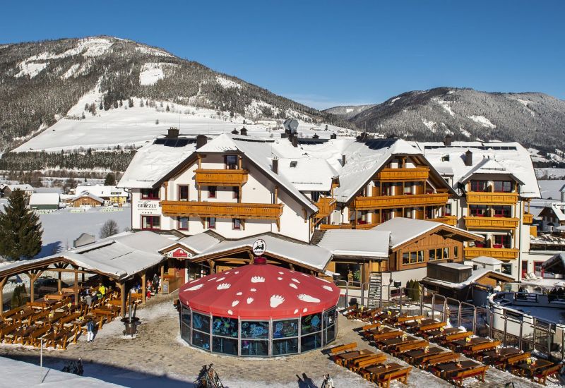 Schneebedecktes Hotel mit Holzfassade im Salzburger Lungau, umgeben von verschneiten Bergen und blauen Himmel.