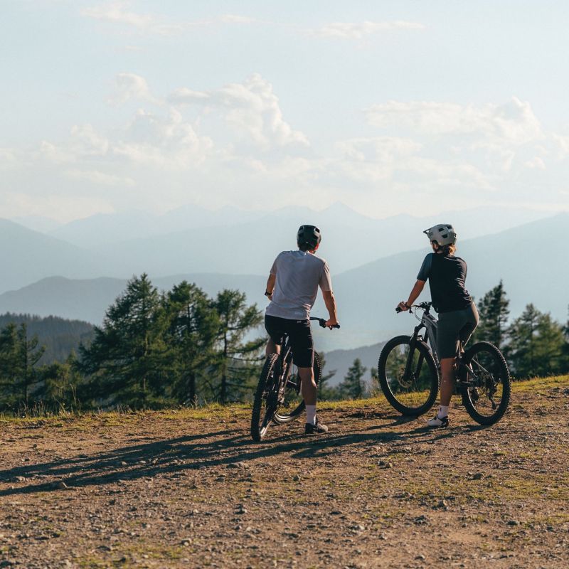 Zwei Radfahrer mit Helmen stehen auf einem Hügel im Salzburger Lungau und blicken auf bewaldete Berge in der Ferne.