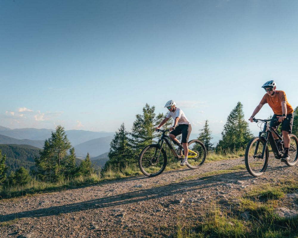 Zwei Personen fahren mit Mountainbikes auf einem Schotterweg durch eine bergige Waldlandschaft im Salzburger Lungau.