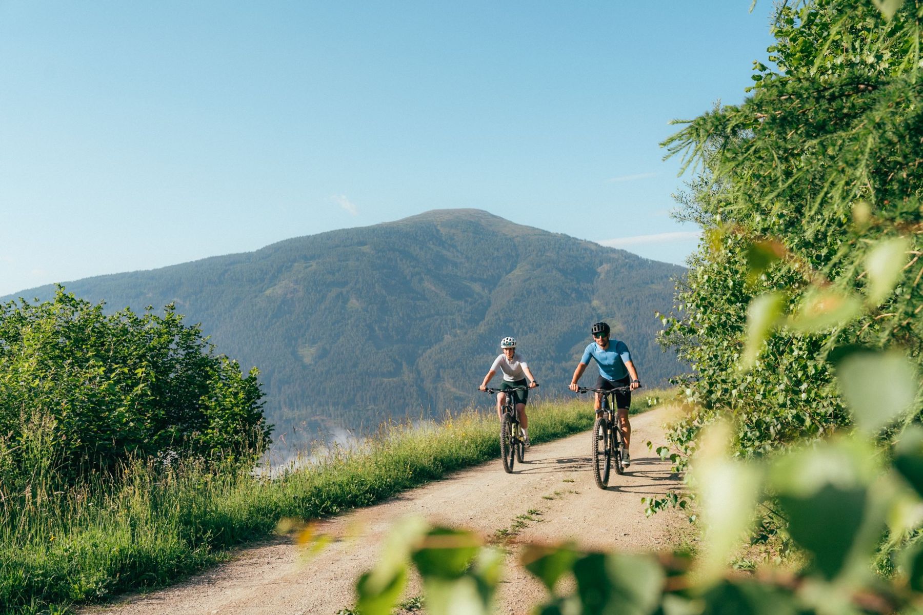 Zwei Radfahrer auf einem Schotterweg in einer grünen, bergigen Landschaft im Salzburger Lungau bei klarem Himmel.