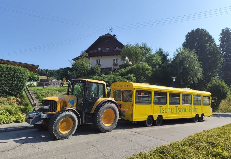 Gelber Traktor zieht gelben Zugwagen mit "Tschu-Tschu-Bahn"-Aufschrift, vor einem Haus im Salzburger Lungau.