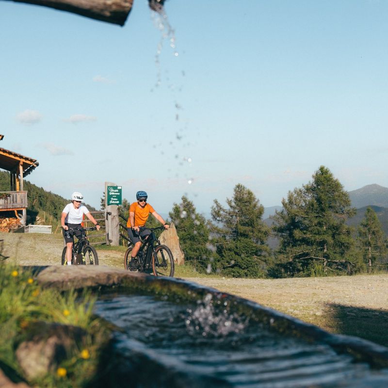 Zwei Radfahrer fahren auf einem Weg neben einer Holzhütte in einer bergigen Landschaft mit Bäumen und einem Holzbrunnen.