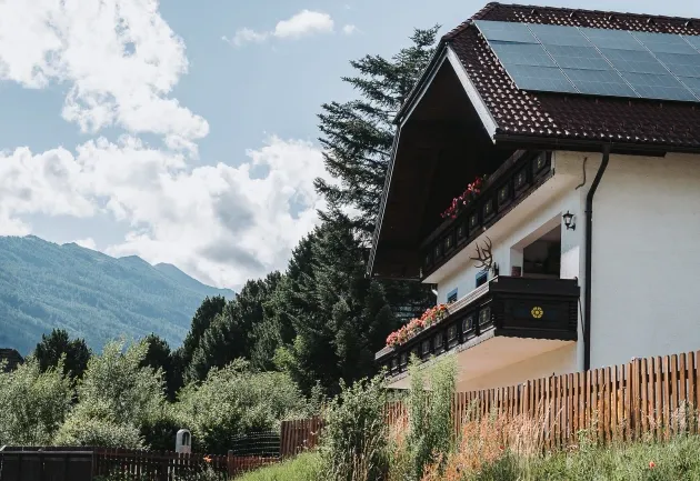 Weißes Haus mit Solarpanelen und Balkon, umgeben von Bäumen und Bergen im Salzburger Lungau, bei sonnigem Wetter.