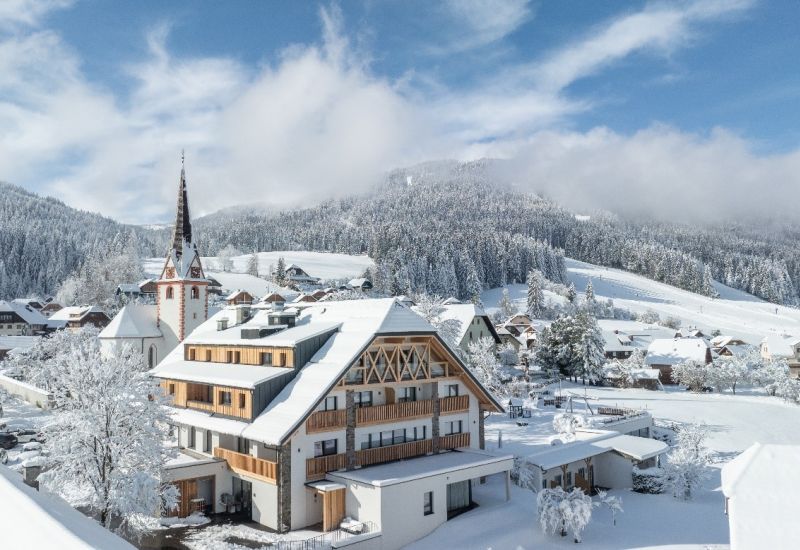 Verschneite Landschaft in Salzburgs Lungau mit Kirche, Gebäuden und bewaldeten Hügeln im Hintergrund unter blauem Himmel.