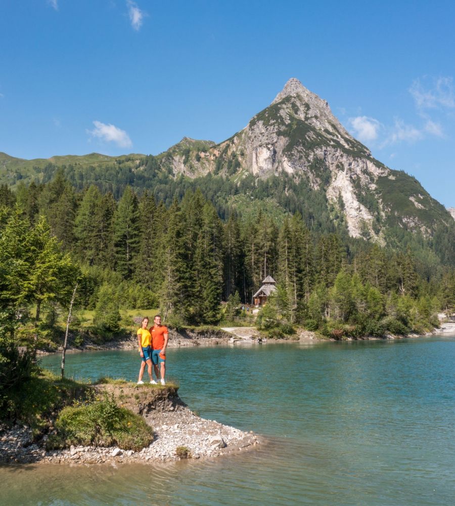 Zwei Personen stehen am Seeufer, umgeben von Wald und Bergen, unter klarem, blauem Himmel im Salzburger Lungau.
