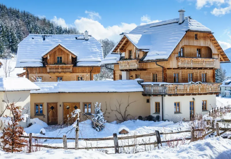 Verschneite Chalets im Salzburger Lungau, umgeben von schneebedeckten Bäumen und blauem Himmel.